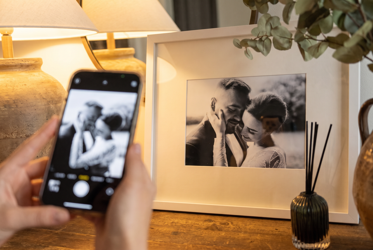 A hand holding a phone in front of a white Framewick frame containing the same black and white wedding photo