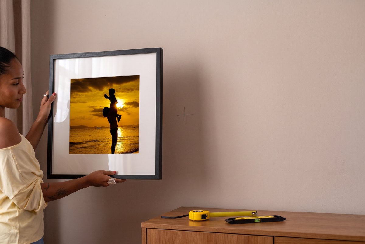 A woman hanging a black Framewick frame with a sunset photo, next to a pencil mark on the wall, with a tape measure and spirit level on the sideboard
